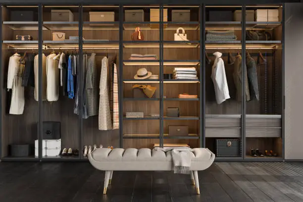 A serene, beautifully organized walk-in closet with soft natural light, neatly hung clothing arranged by color, open shelving with folded items, and a plush bench at the center